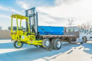 A bright yellow truck-mounted forklift attached to the back of a flatbed delivery truck, parked outdoors on a sunny day, with a blue container secured on the flatbed and industrial power lines visible in the background.